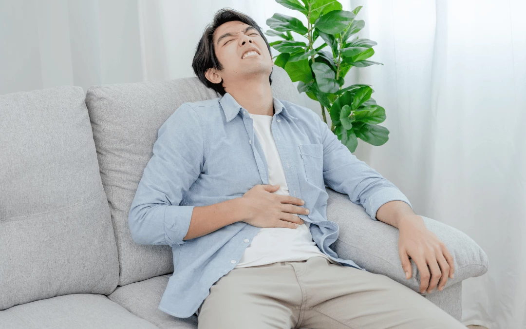 A Man Sitting On Grey Sofa In Pain With His Hand On His Stomach